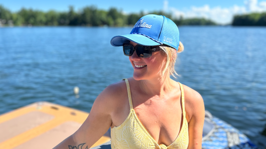 A smiling woman on a boat wearing a blue mesh baseball cap with white script lettering, black sunglasses, and a pale yellow bikini top, with water and trees in the background.