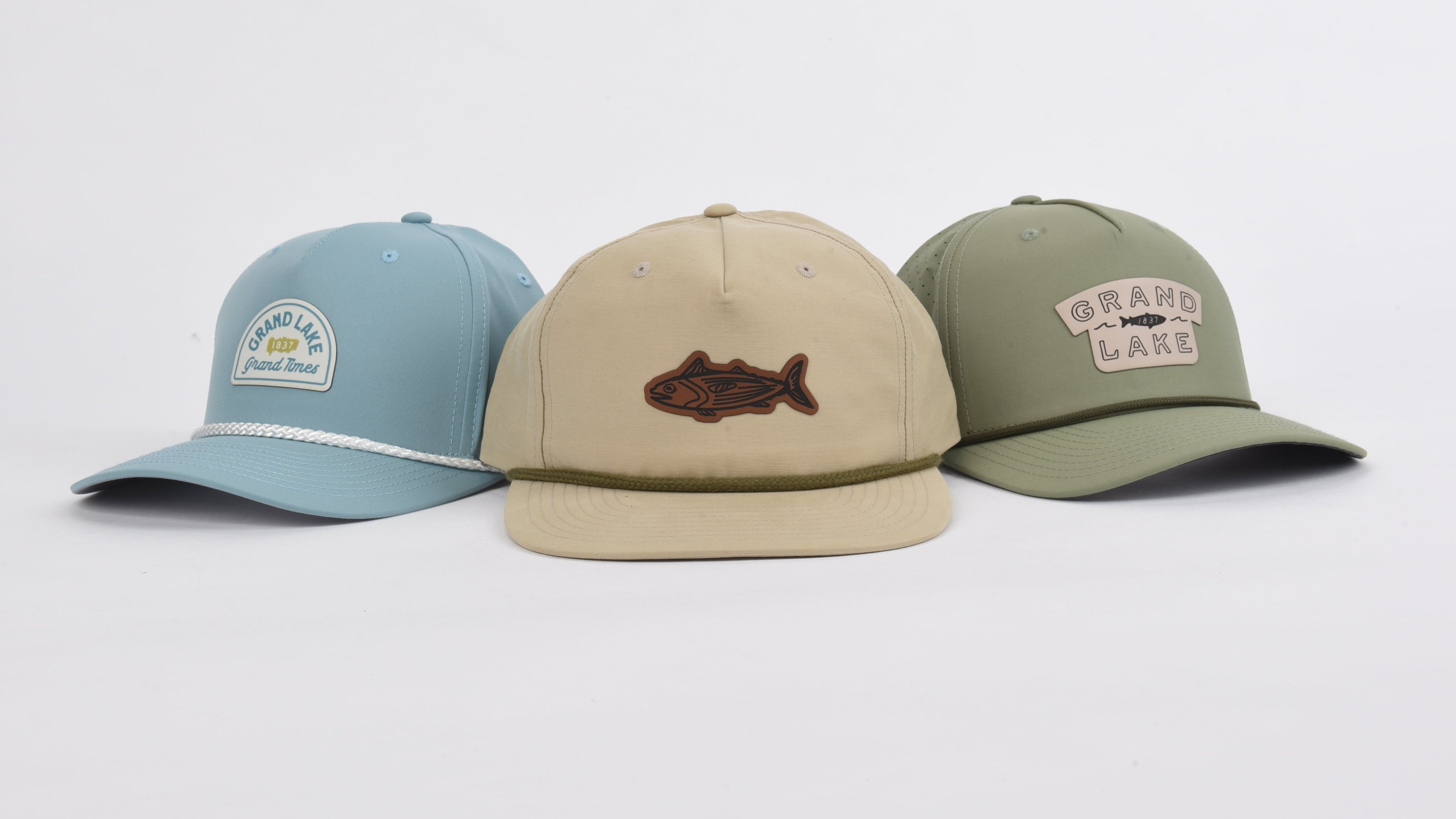 Three flat-brim baseball caps displayed side by side on a light gray background: a light blue cap with a white embroidered patch, a beige cap with a brown fish patch, and an olive green cap with a beige 'Grand Marsh' patch.