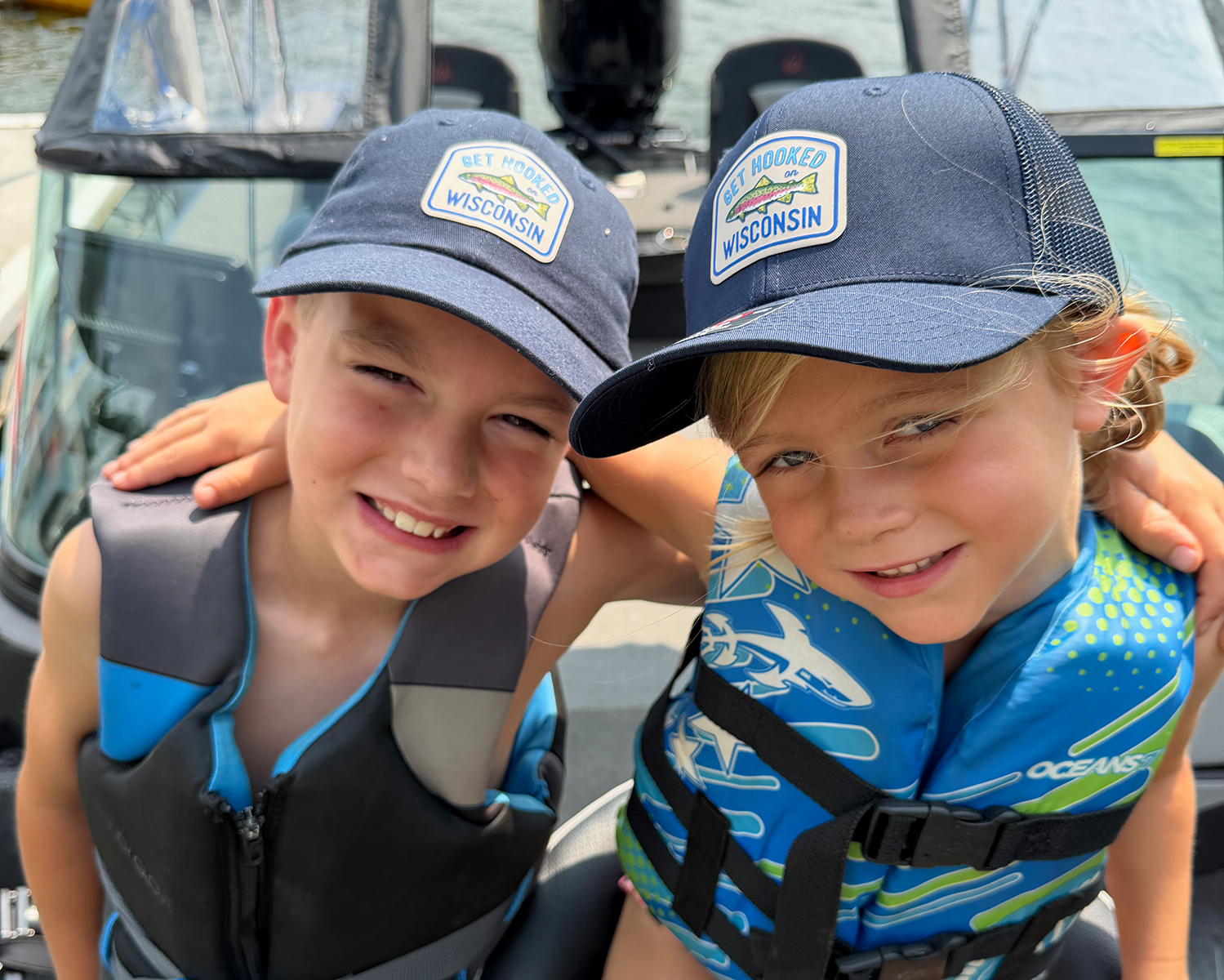 Two children smiling and posing together outdoors, wearing matching blue mesh caps with a 'Wisconsin' patch and blue life vests.