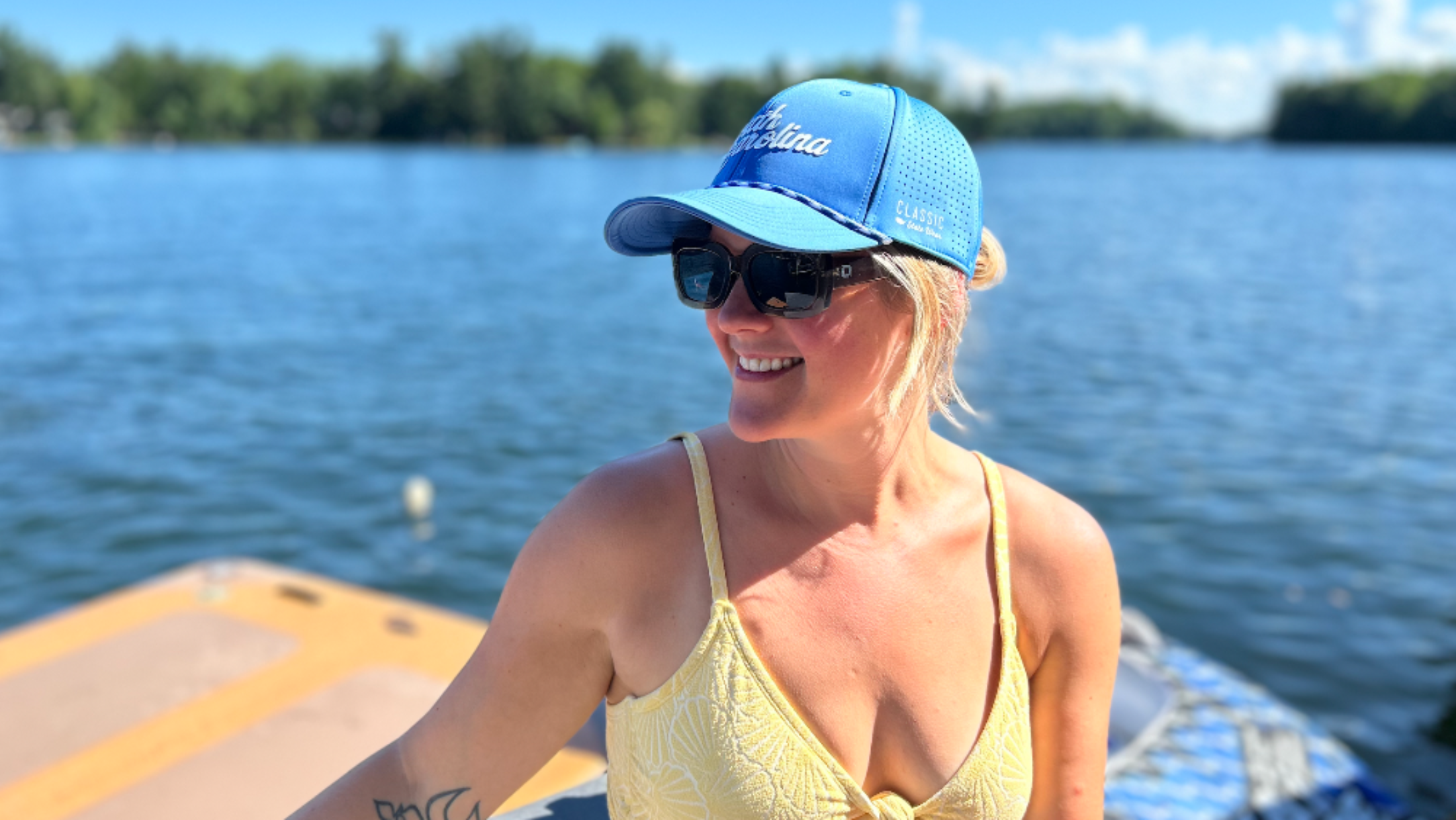 A smiling woman on a boat wearing a blue mesh baseball cap with white script lettering, black sunglasses, and a pale yellow bikini top, with water and trees in the background.